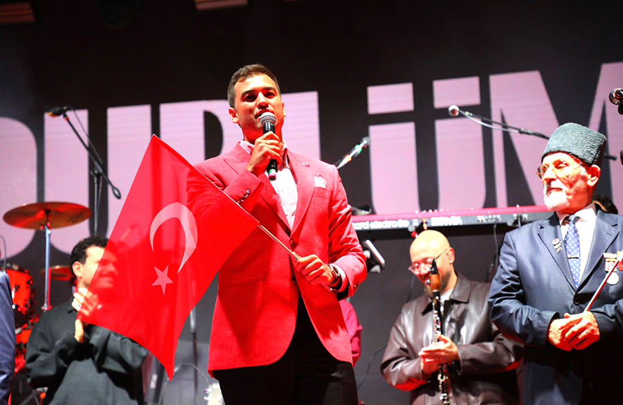 Elinde Türk bayrağı ve mikrofon tutan kırmızı ceketli adam.
