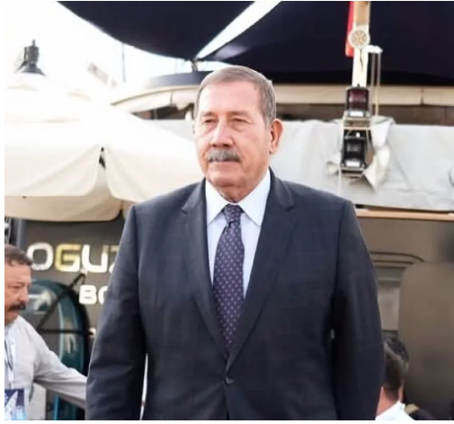 Koyu takım elbiseli, bıyıklı ve ciddi ifadeli yaşlı adam.