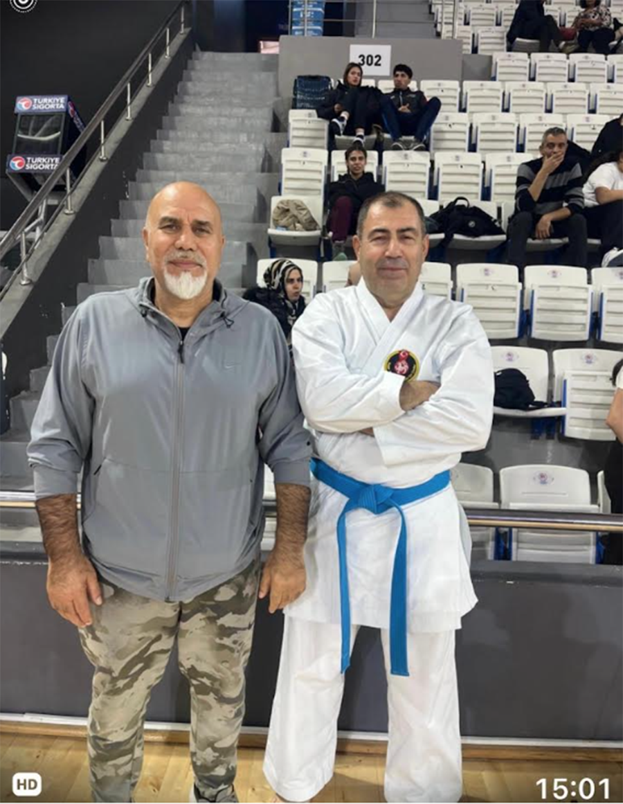 Tribünlerin önünde poz veren bir antrenör ve mavi kuşaklı karateci.