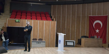 Kürsü ve Türk bayrağı bulunan sahnede duran bir adam.