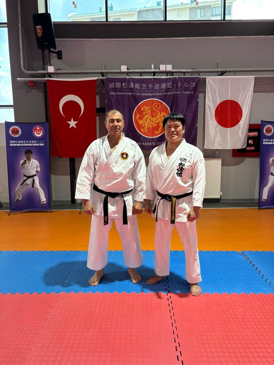 Karate kıyafetli ve siyah kuşaklı iki usta bayraklar önünde.